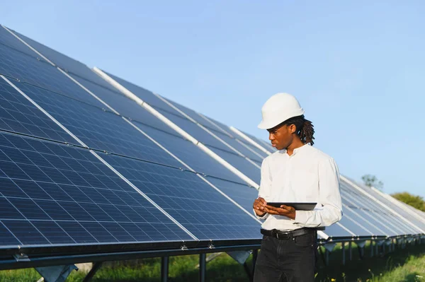 Büyük güneş panellerini denetleyen, optimal performans ve sürdürülebilir enerji üretimini sağlayan, sert şapkalı Afrikalı Amerikalı bir adam.