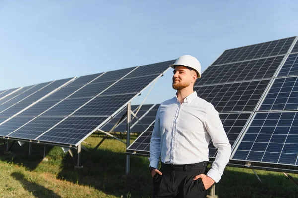 Sert şapkalı ve gömlekli bir adam büyük bir güneş panelinin önünde gururla duruyor, temiz enerjiyi ve sürdürülebilir geleceği temsil ediyor.
