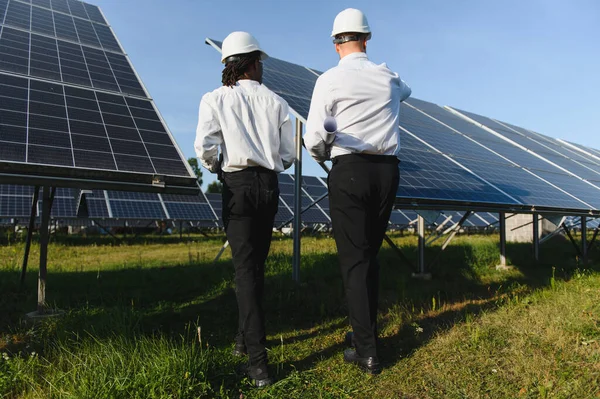 Mühendisler açık bir gökyüzü altında sürdürülebilir enerji üretimine odaklanmış büyük bir güneş çiftliğindeki güneş panellerini aktif olarak kontrol ediyorlar.