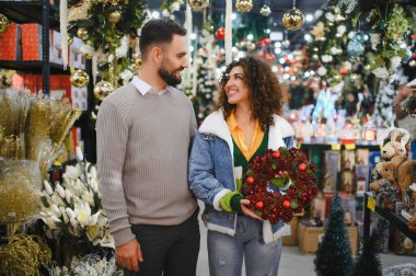 Mutlu çift, bayram süsleriyle dolu bir mağazada birbirlerine bakarken kırmızı bir çelenk seçiyorlar.