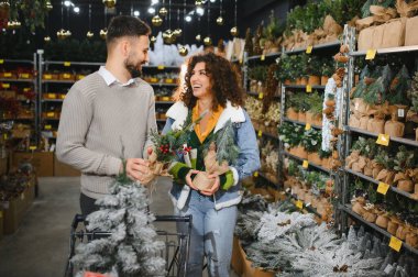 Mutlu genç çift bayram süslemelerini seçiyor, ellerinde minyatür ağaçlar ve mağazada gülümsüyor.