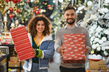 Noel mağazasında gülümseyen genç bir çift, ellerinde parlak kırmızı hediye kutuları, birlikte tatil hediyeleri alırken, neşeli alıcılar