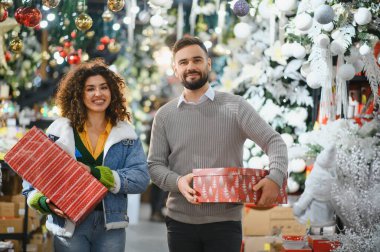Genç çift gülümsüyor, hediye paketliyor ve Noel süsleri ve süslemelerle dolu bir mağazada yürüyor.