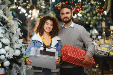 Gülümseyen genç çift ellerinde hediye kutuları, süslü ağaçlar ve ışıklarla bayram alışverişi yapıyorlar.