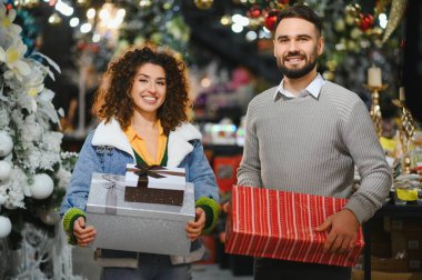 Genç bir çift hediye taşıyor, kutlama dekorasyonları önünde kameraya gülümsüyor. Tatil sezonunu kutluyoruz
