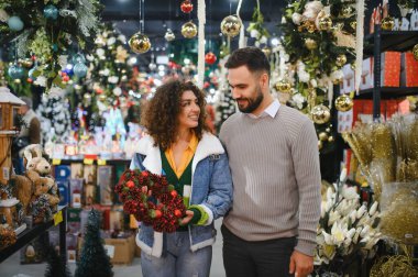 Neşeli bir çelenk seçen mutlu çift, ışıltılı süslemelerle dolu bir mağazada tatil sezonunun tadını çıkarıyor.