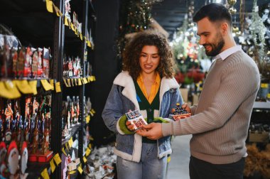 Gülümseyen genç çift bayram dekorlarını seçiyor, Noel ışıkları olan bir mağazada minyatür evleri tutuyorlar.