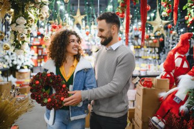 Noel çelengi seçen mutlu çift Noel süsleri ve hediyelerle dolu bir mağazadan Noel alışverişi yapıyor.