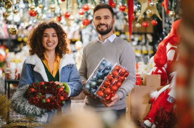 Genç çift gülümsüyor, ellerinde Noel süsleri ve çelenkler var, bayram dekoru alışverişinin tadını çıkarıyorlar. Şenlikli perakende deneyimi