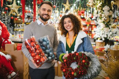 Genç adam ve kadın gülümsüyor, ellerinde bayram süsleri ve dekoratif bir çelenk var, festival sezonuna hazırlanıyorlar.