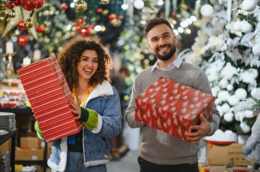Gülümseyen genç bir çift ellerinde hediye kutularıyla bayram süslemeleriyle bayram mağazasında Noel alışverişinin tadını çıkarıyor.
