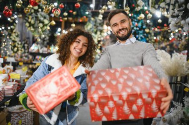 Genç çift gülümsüyor, ellerinde süslü hediyeler var. Parlak ışıklı mevsimlik bir mağazada bayram alışverişi yapıyorlar.