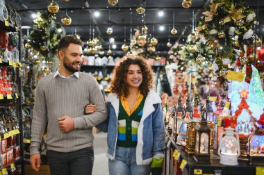 Gülümseyen ve kol kola yürüyen bir çift gelecek Noel tatili için şenlikli süslemeler seçiyor.
