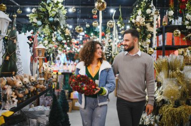 Genç çift birbirine bakıyor, gülümsüyor ve bayram süsleri ve şenlik eşyalarıyla dolu bir mağazada yürüyor, çelenk alıyorlar.