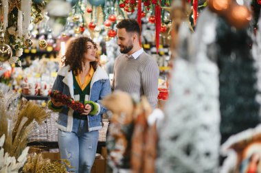 Genç çift gülümserken parlak bir bayram dekoru mağazasında Noel süsleri ve çelenkler seçiyor, rahat mevsimlik alışveriş sahneleri.