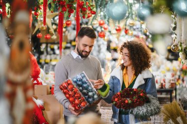 Mutlu genç çift neşeli bir bayram pazarında renkli Noel süsleri ve çelenkler seçiyor, gülümsüyor ve ev dekorasyonu hazırlıyorlar.