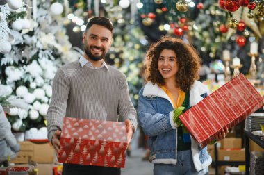 Genç çift kameraya bakıyor, ellerinde hediyeler ve bayram süsleme mağazasında gülümsüyor.