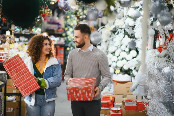 Genç çift bir mağaza koridorunda yürüyor, bayram döneminde Noel hediyeleri ve dekorasyonlar alıyorlar.