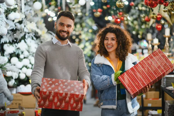 Genç çift kameraya bakıyor, ellerinde hediyeler ve bayram süsleme mağazasında gülümsüyor.