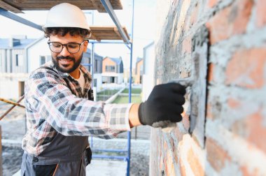 İnşaat alanında yeni bir tuğla duvar ören sert bir şapka ve koruyucu eldiven takan yetenekli bir inşaat işçisi.