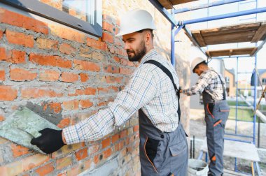 Duvara havan topu uygulayan, sıkı şapka takan, üniformalı bir inşaat işçisi. Yeni ev inşa etmek ya da eskisini yenilemek.