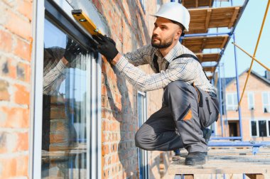 Sert şapka ve iş üniforması giyen inşaat işçisi, düz bir araçla tuğla binanın dış tarafına yeni bir pencere kuruyor.