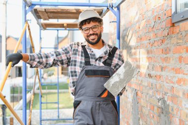 Erkek işçi, kasketli ve tulumlu, havan topu taşıyan bir mala tutuyor, inşaat alanında iskelenin yanında duruyor.