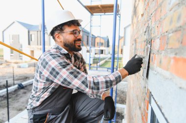 İnşaat işçisi, sert şapka ve ekose gömlek giyip inşaat alanındaki bir malayla tuğla duvara havan topu atıyor.