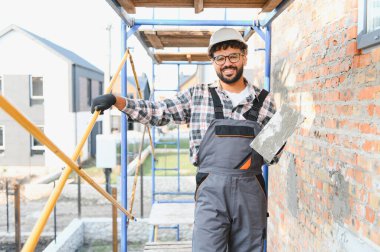 Kasketli ve iş tulumlu bir adam inşaat alanında gülümsüyor, havan topu tutuyor, tuğla döşemeye hazır.