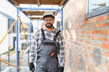 Genç müteahhit kasketli ve tulumlu, tuğla bir duvarın yanında duruyor ve inşaat sahasında iskele yapıyor.