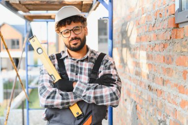 Kasketli, ekose gömlekli ve eldivenli genç adam inşaat alanında tuğla duvara yaslanmış duruyor.