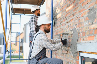İnşaatçılar sert şapkalar ve iş kıyafetleri giyip dış tuğla duvara sıva yapıştırıyor ve bir evi yeniliyorlar.
