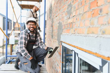 İnşaat işçisi, iskeledeki tuğladan duvara sıva uygularken mutluydu. Arka planda çalışan bir meslektaşı vardı.