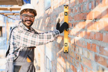 Mutlu inşaat işçisi bir tuğla duvarın hizasını ruh seviyesiyle kontrol ederek geleceği inşa ediyor.