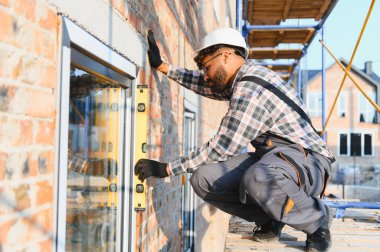 İnşaat işçisi, bir tuğla evin ön cephesine pencere yerleştiriyor. Tam hizalama için ruh seviyesini kullanıyor. Yeni ev inşa ediliyor