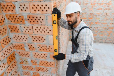 İnşaat işçisi, iş üniforması ve şapka takıyor. İnşaat alanındaki ruhsal seviyeyle tuğla bir duvarın dikey hizasını kontrol ediyor.