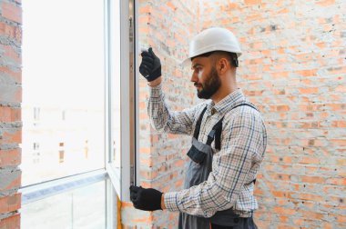 İnşaat işçisi, başlık ve tulum giyerek tuğla duvar açılışına yeni pencere çerçevesi yerleştiriyor ve evi yeniliyor.