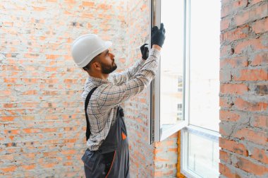 İnşaat işçisi yeni evine pencere paneli yerleştiriyor. Kasketi ve koruyucu eldivenleri var. Yenileme ve inşa konsepti