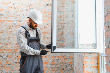 İnşaatçı, inşaat halindeki bir binaya turuncu ve gri üniformalı beyaz başlıklı yeni bir plastik pencere takıyor.