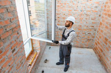 İnşaat işçisi bir inşaat sahasına modern bir pvc pencere çerçevesi kuruyor, tuğla bir duvar üzerinde çalışıyor.