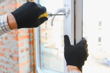 İşçi, ev tadilatı sırasında beyaz plastik çerçeveye bir tornavidayla pencere sapını takan eldivenler takıyor.