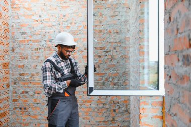 Afrikalı Amerikalı inşaatçı tamamlanmamış tuğla bir duvara yeni bir pvc pencere çerçevesi yerleştiriyor. Ev yenileme projesi