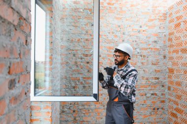Başlık ve tulum takan genç adam tamamlanmamış tuğla bir duvara yeni bir pencere çerçevesi yerleştiriyor.