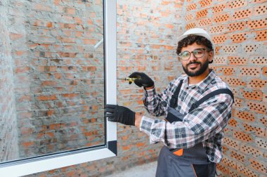 İnşaat işçisi bir pencere kuruyor, bir tornavida tutuyor, inşaat alanında kurulum yapıyor.