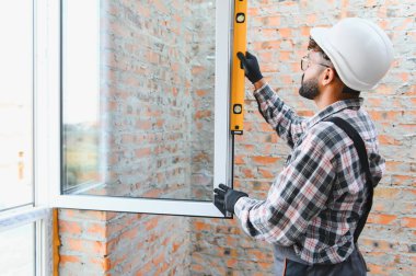 Kalın kasklı inşaat işçisi tuğla duvar açılışında metal pencere çerçeveleri kuruyor, seviye hizalaması sağlanıyor