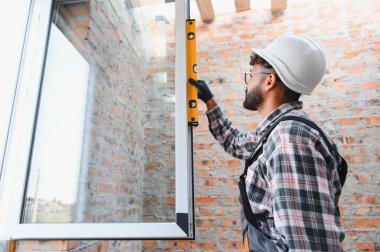 Sert şapka takan ve iş kıyafetleri giyen bir adam binadaki yeni pencere kurulumunu doğru ölçüyordu. Ev yenileme ve iyileştirme