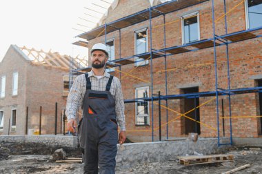 İnşaatçı inşaat halindeki yeni tuğla evi teftiş ediyor, sıkı bir şapka ve tulum giyiyor. Gayrimenkul geliştirme