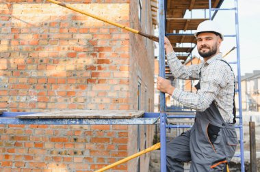 İnşaat alanındaki metal iskeleye tırmanan kasketli ve tulumlu işçi kameraya bakıyor.
