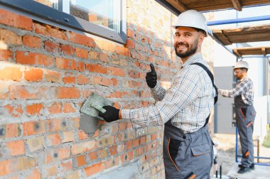 İnşaat işçisi, başlık ve üniforma takıyor, kameraya gülümsüyor, duvarları sıva yaparken başparmağını kaldırıyor.