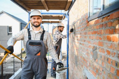 Profesyonel müteahhit, inşaat iskelesinde üniforma takıyor, tuğla duvar yapıştıran başka bir işçinin yanında mala tutuyor.